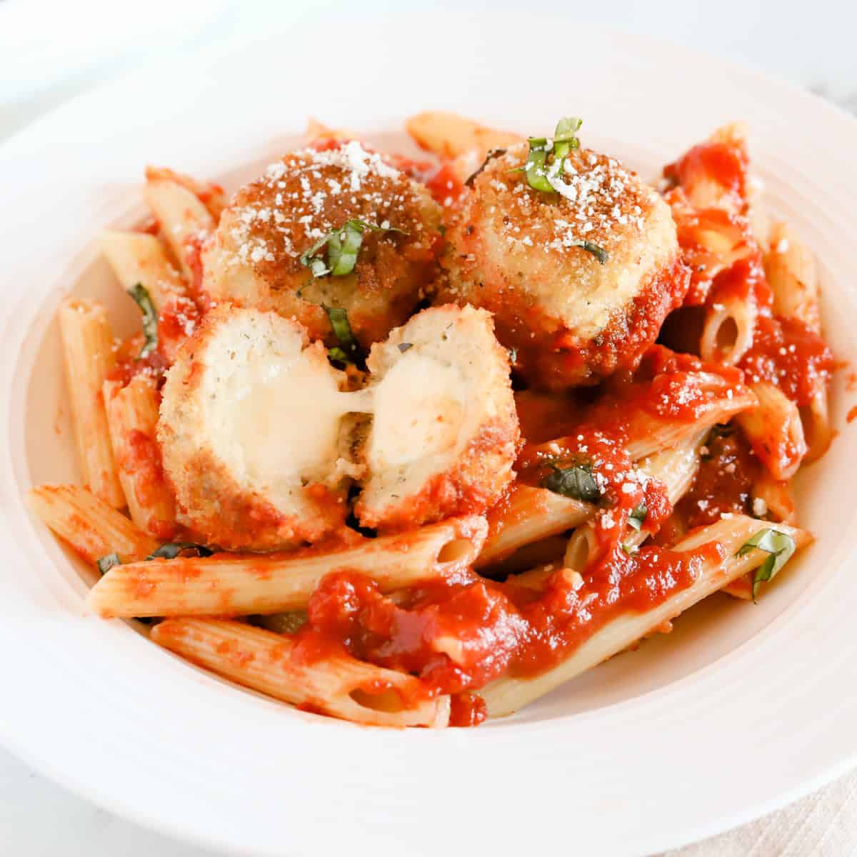Chicken parmesan meatballs served with penne noodles. One meatball is cut in half exposing the melted mozzarella.