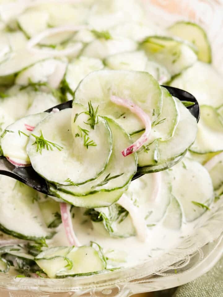 black spoon with a scoop of cucumber dill salad from a glass serving bowl.