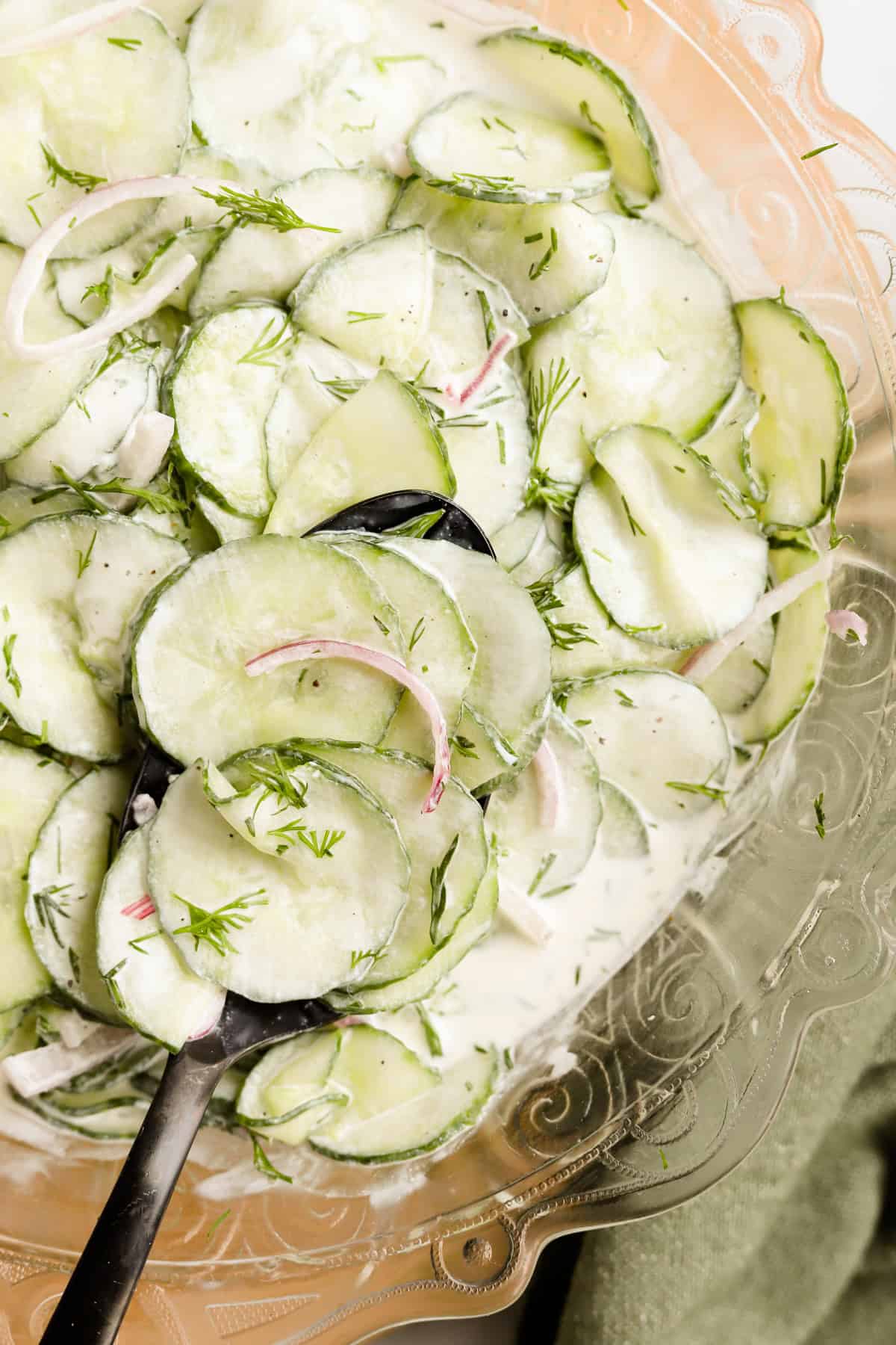 Overhead view of cucumber dill salad in a glass serving bowl with a black spoon.