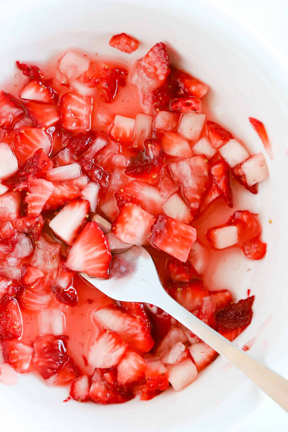 Chopped strawberries macerating in a white bowl with a fork smashing them