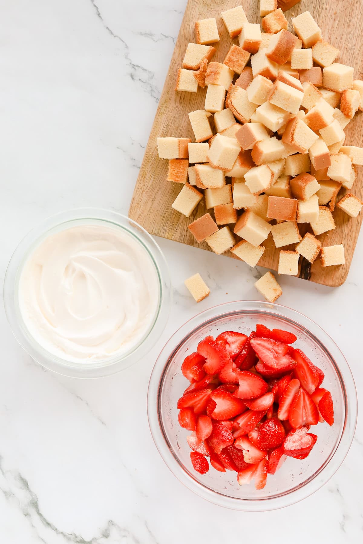 Wooden cutting board of pound cake alongside a bowl of macerated strawberries and whipped cream.