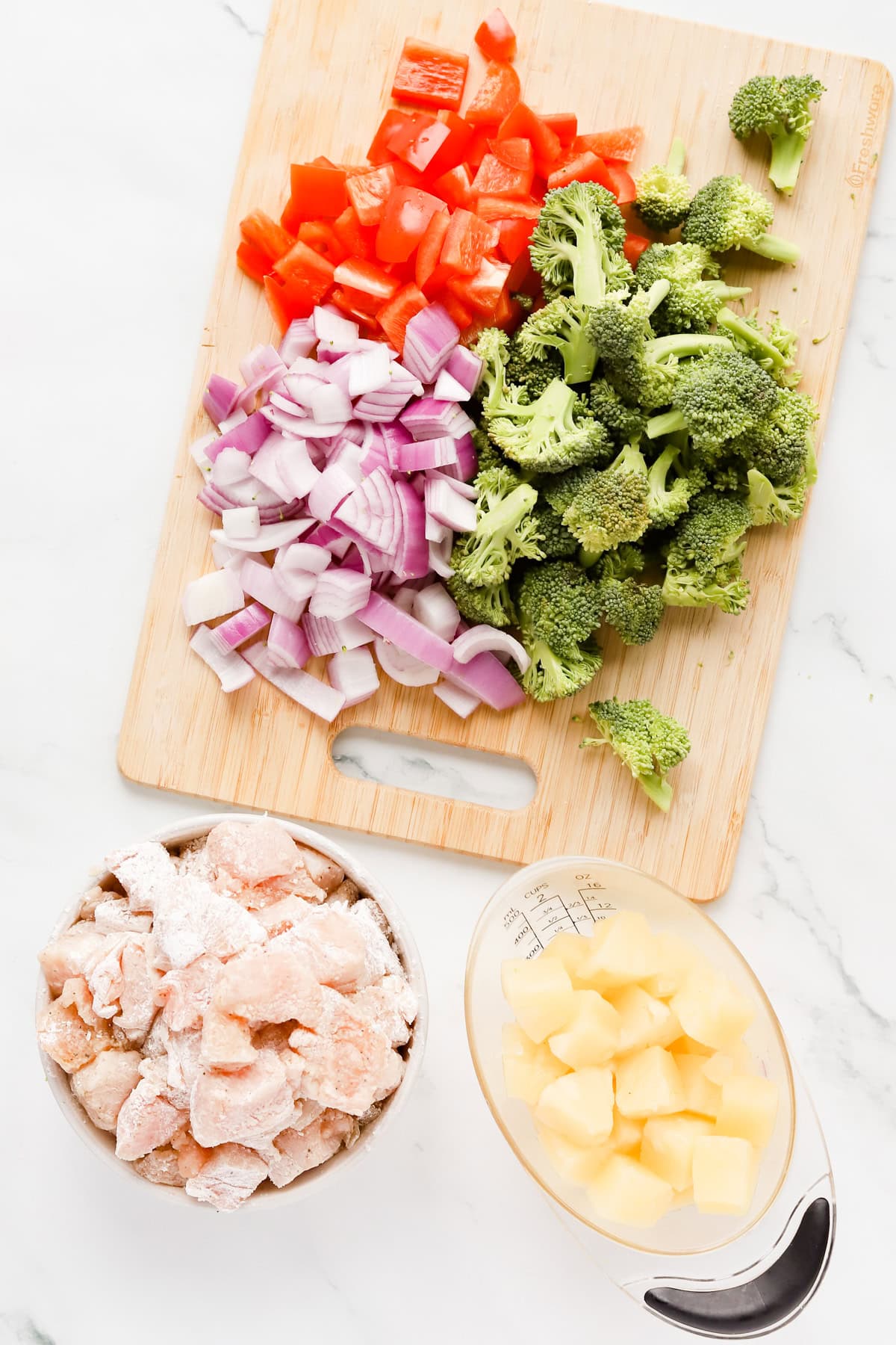 Chopped broccoli, red pepper and red onion a wooden cutting board alongside a bowl of cornstarch coated chicken pieces and clear measuring cup of pineapple chunks.