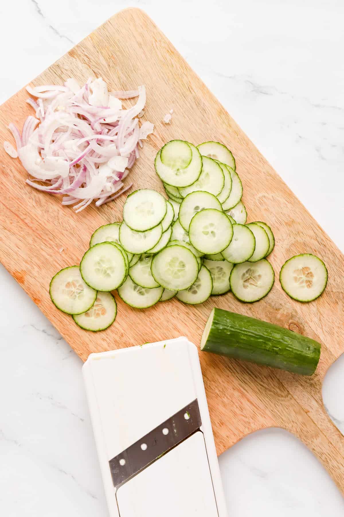 Overhead view of sliced red onion and cucumber with a mandoline on a wooden cutting board.