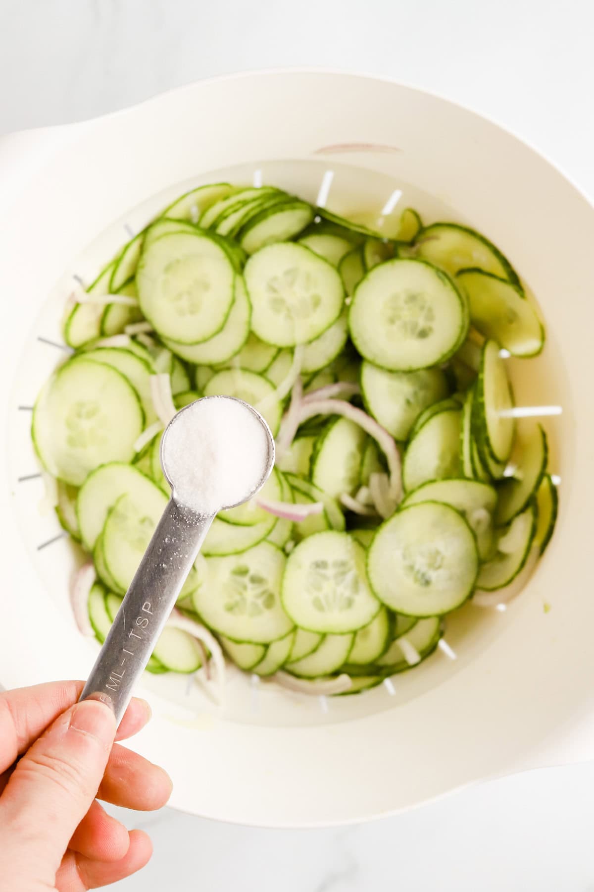 A teaspoon of salt ready to be sprinkled on sliced cucumbers in a white colander.