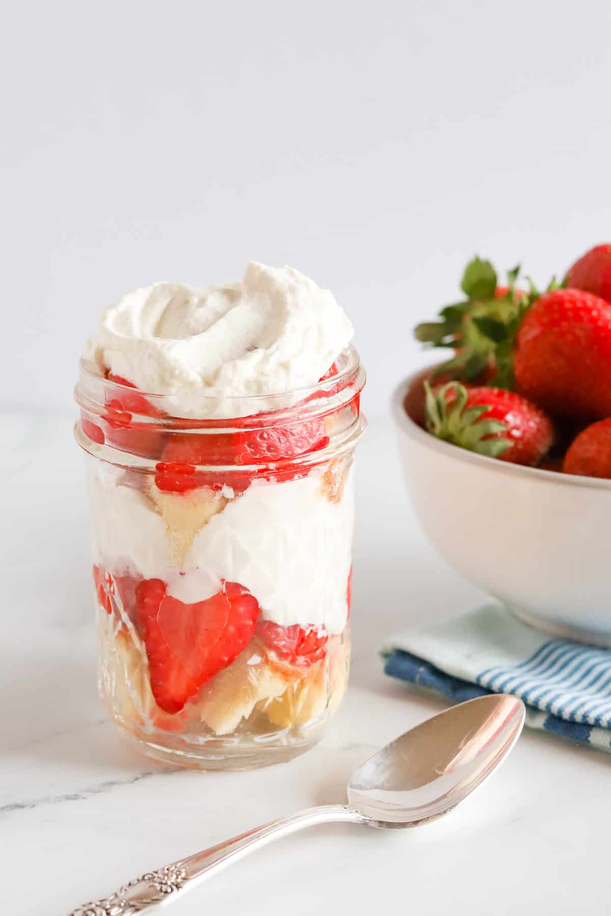 Assembled strawberry shortcake parfaits with a silver spoon and bowl of strawberries next to it.