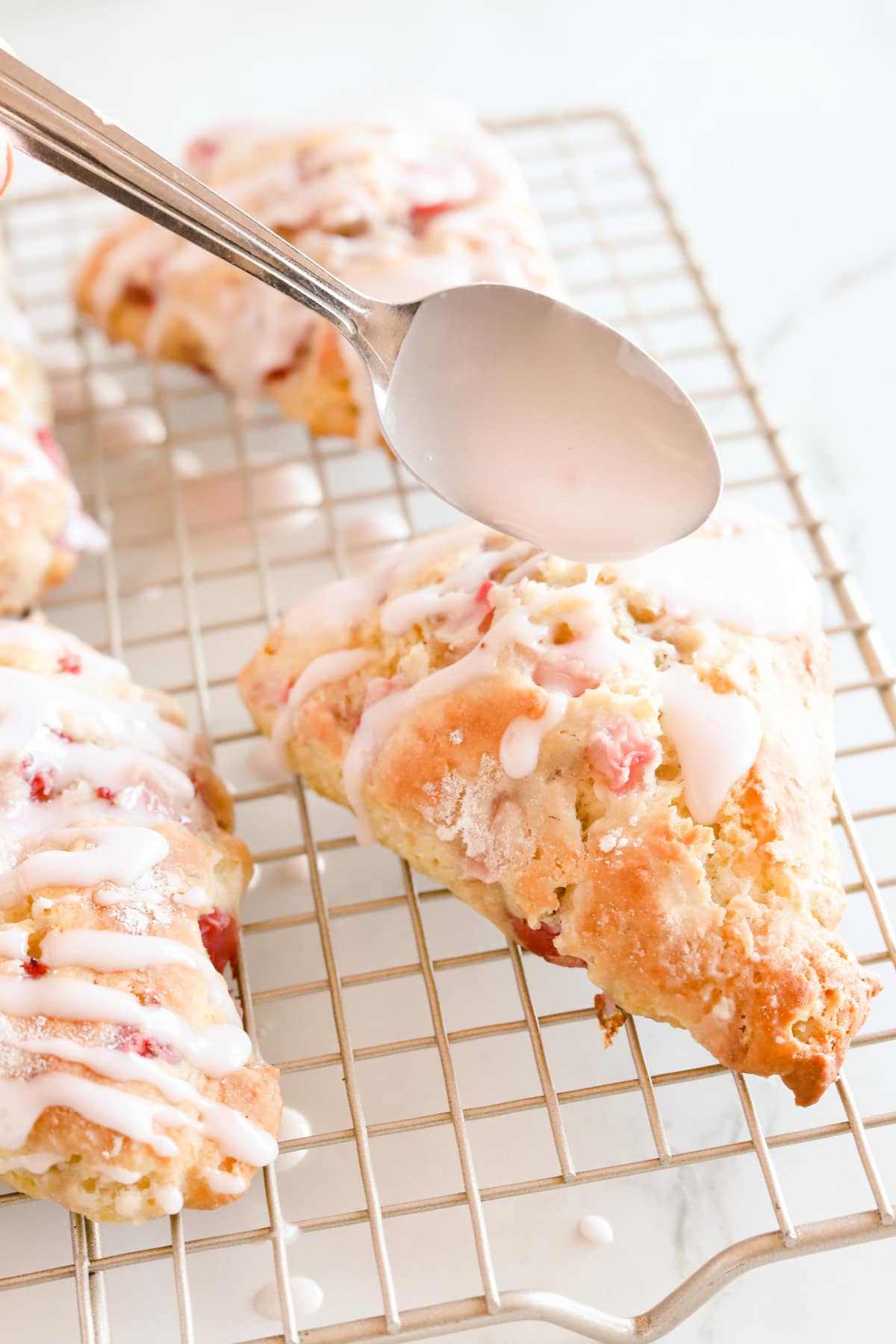 Spoon drizzling strawberry glaze over the top of scones on a wire rack.