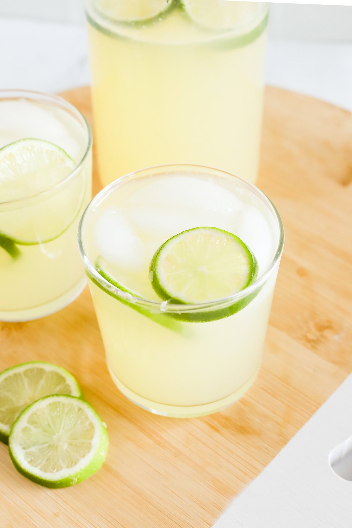 Limeade in two glasses with ice and lime slices on a wooden tray.