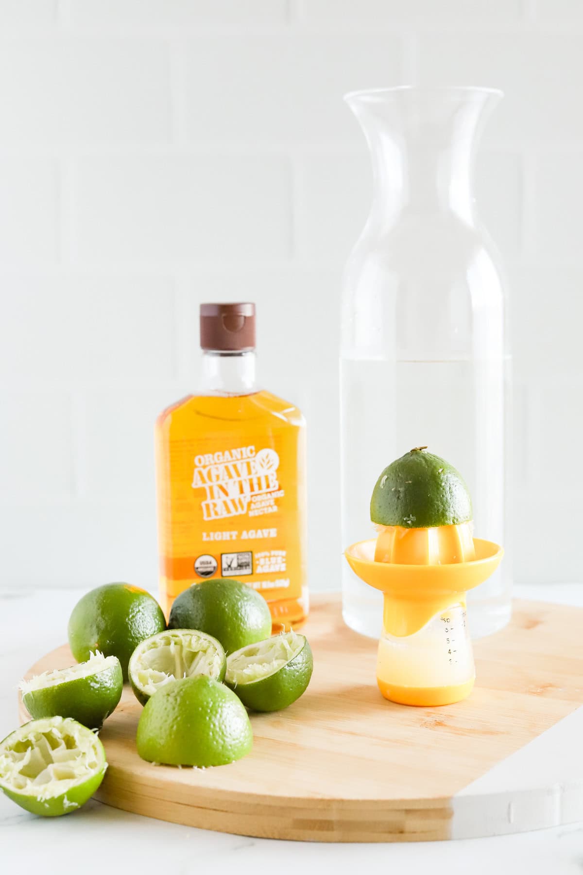 Citrus juicer, carafe of water, juiced limes and bottle of agave nectar on a round wooden tray.