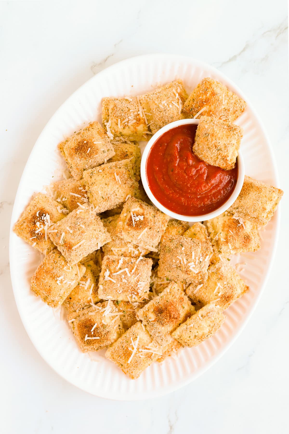 Crispy baked toasted ravioli on a white oval platter with a white bowl of marinara sauce.