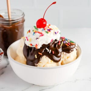 Featured image of a hot fudge sundae in a white bowl with rainbow sprinkles, whipped cream and a cherry on top.