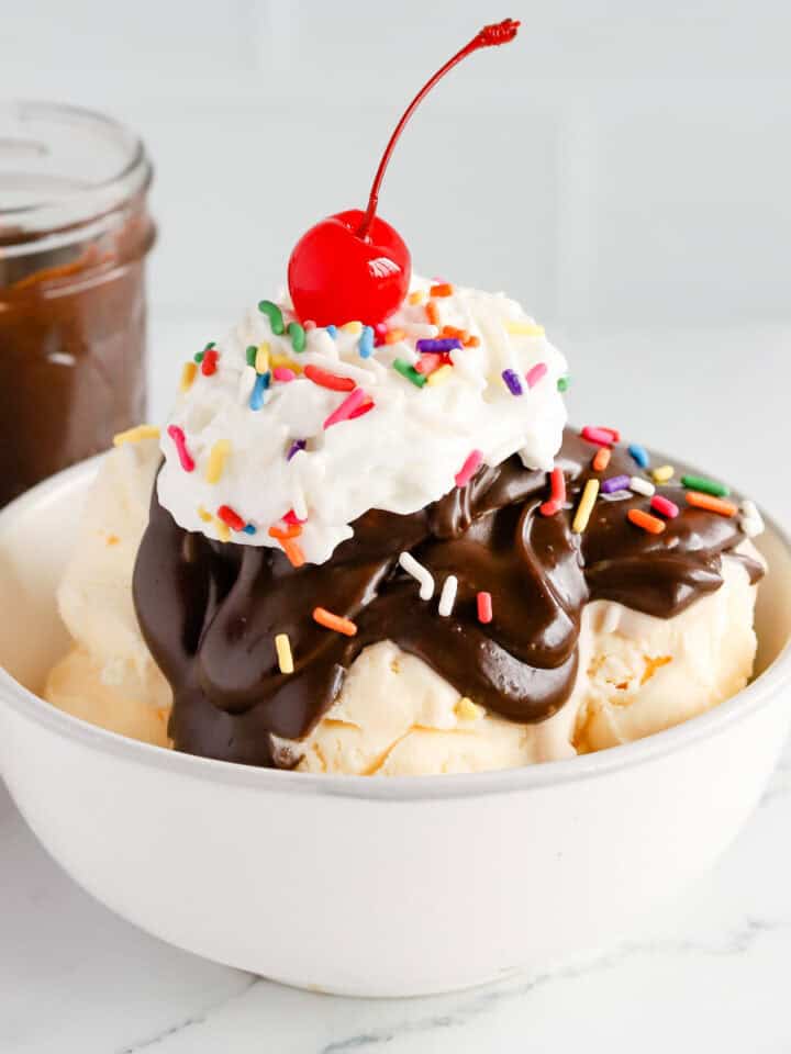 Featured image of a hot fudge sundae in a white bowl with rainbow sprinkles, whipped cream and a cherry on top.