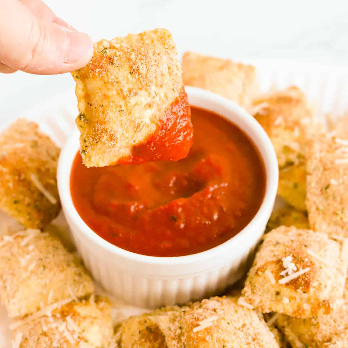 Hand dipping a toasted ravioli in a white bowl of marinara.
