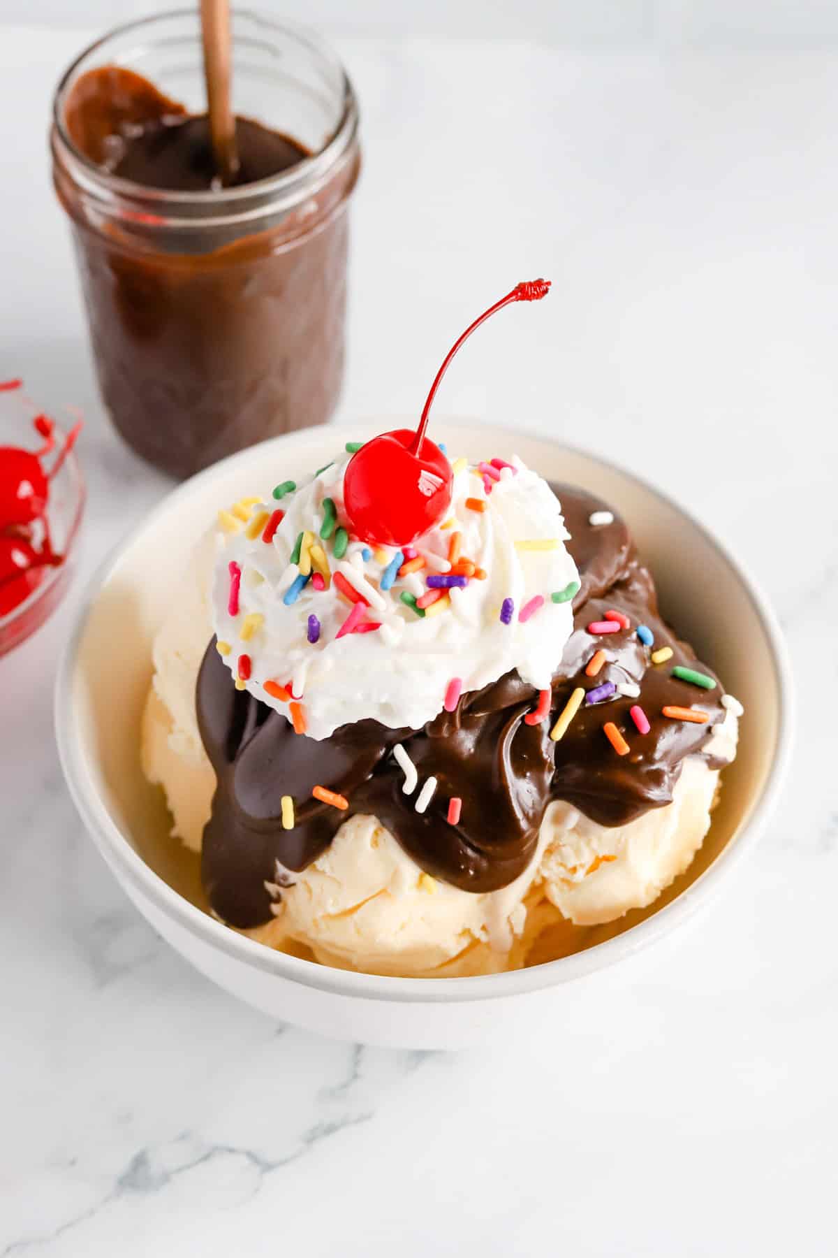 Hot fudge sundae in a white bowl with a jar of hot fudge and cherries in a bowl behind it.