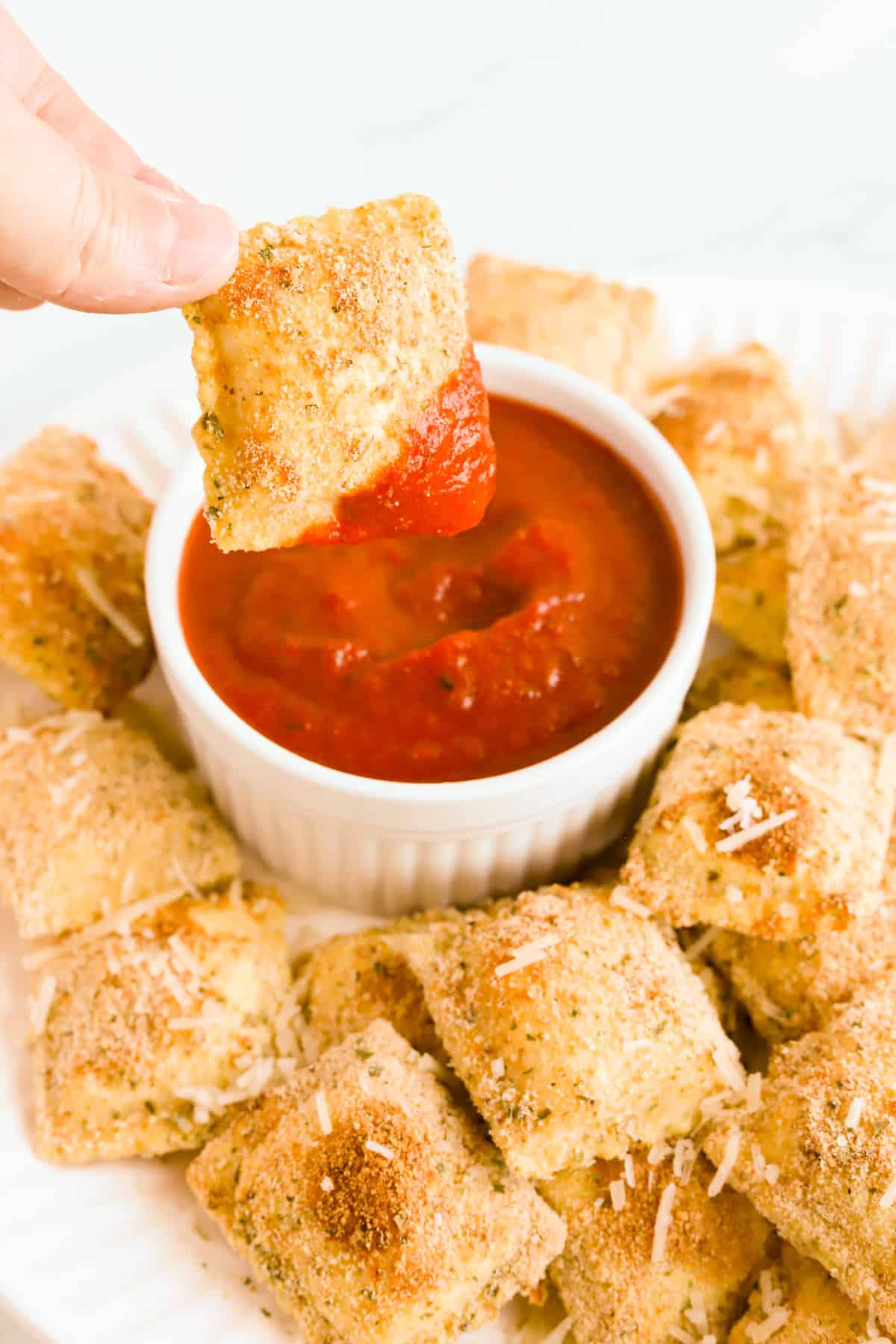Hand dipping a toasted ravioli in white bowl of marinara on a platter of ravioli.