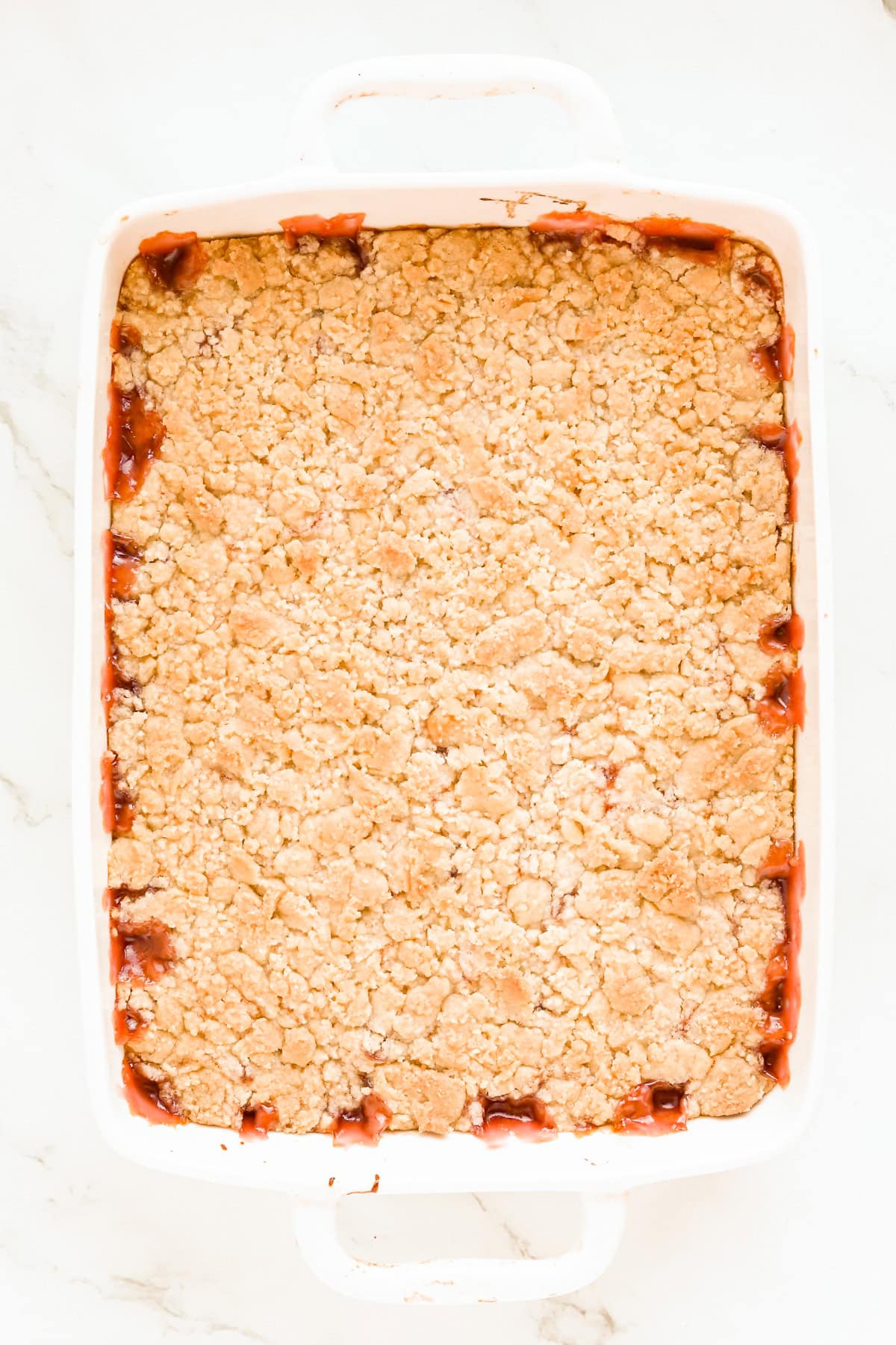 Baked cherry cobbler in a white 9x13 baking dish. The topping is golden brown and some cherry filling is leaking around the edges.