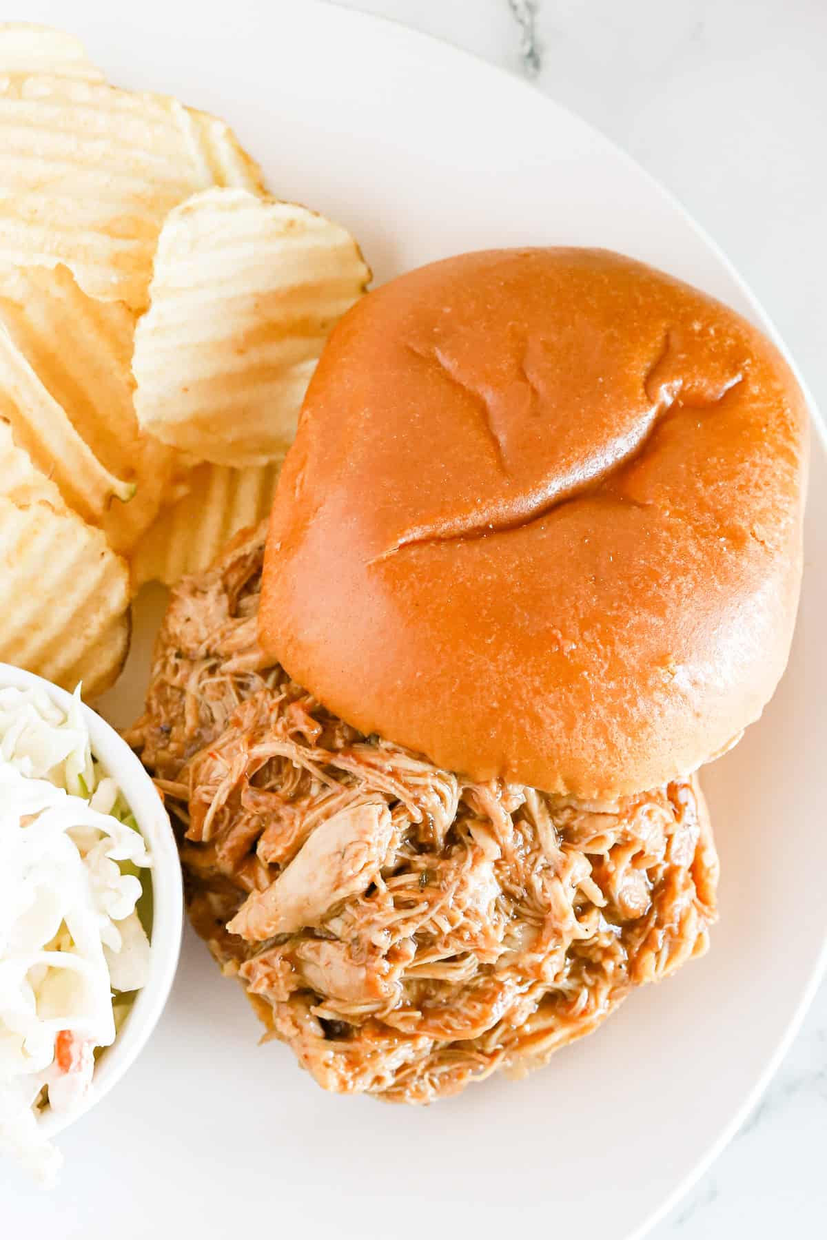 Overhead shot of bbq chicken sandwich on a white plate with coleslaw and potato chips.