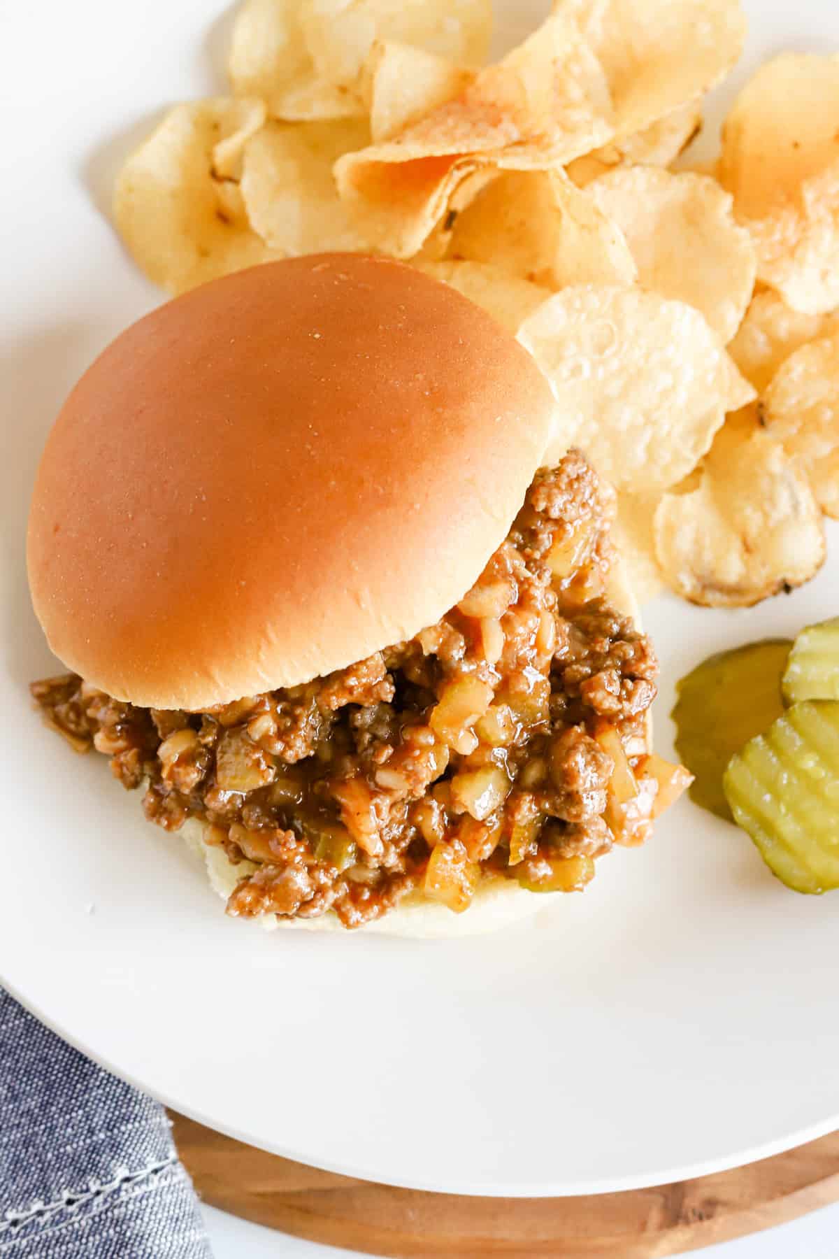 Overhead of a sloppy Joe sandwich with potato chips and pickle slices on a white plate.