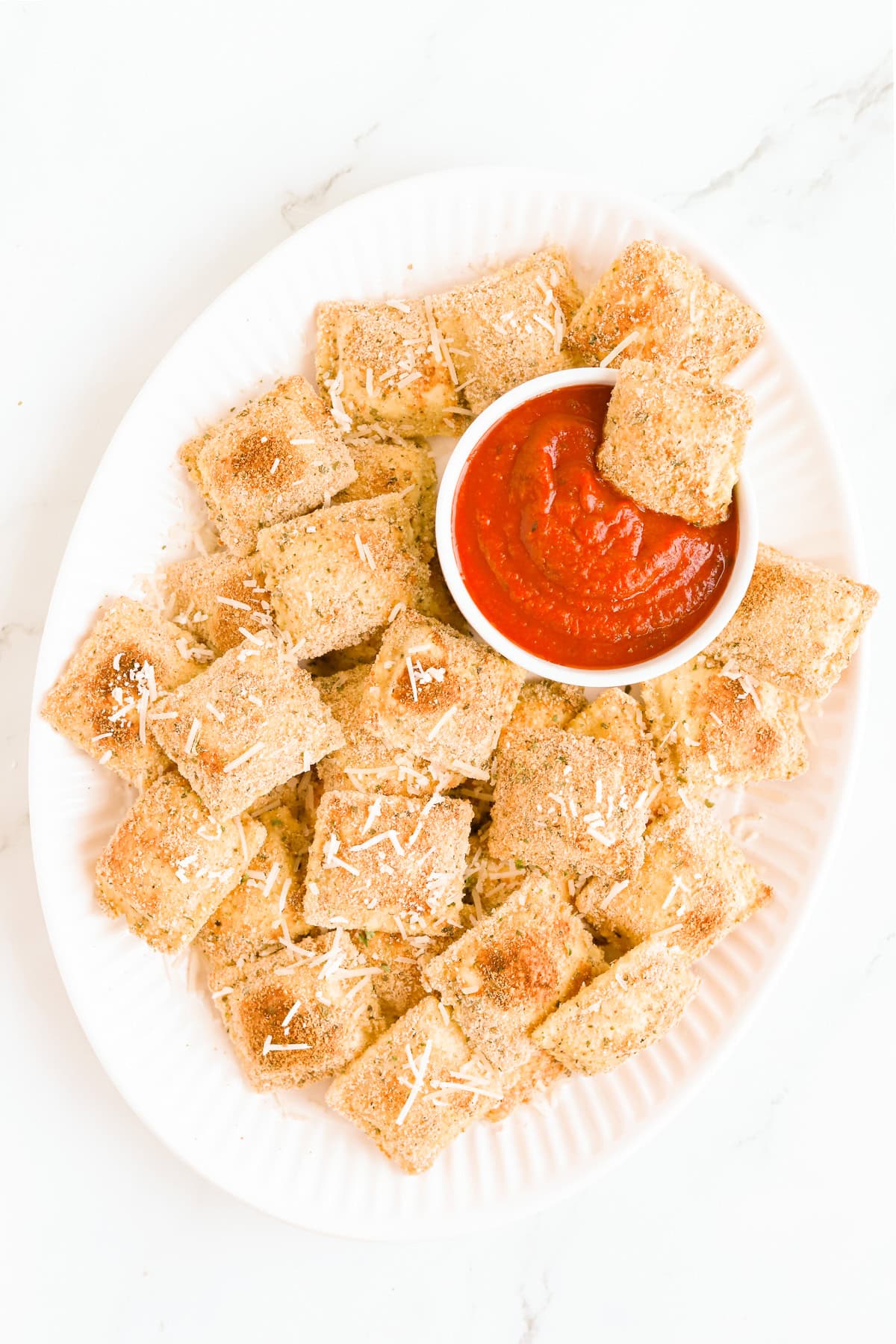 Crispy baked toasted ravioli on a white oval platter with a white bowl of marinara sauce.