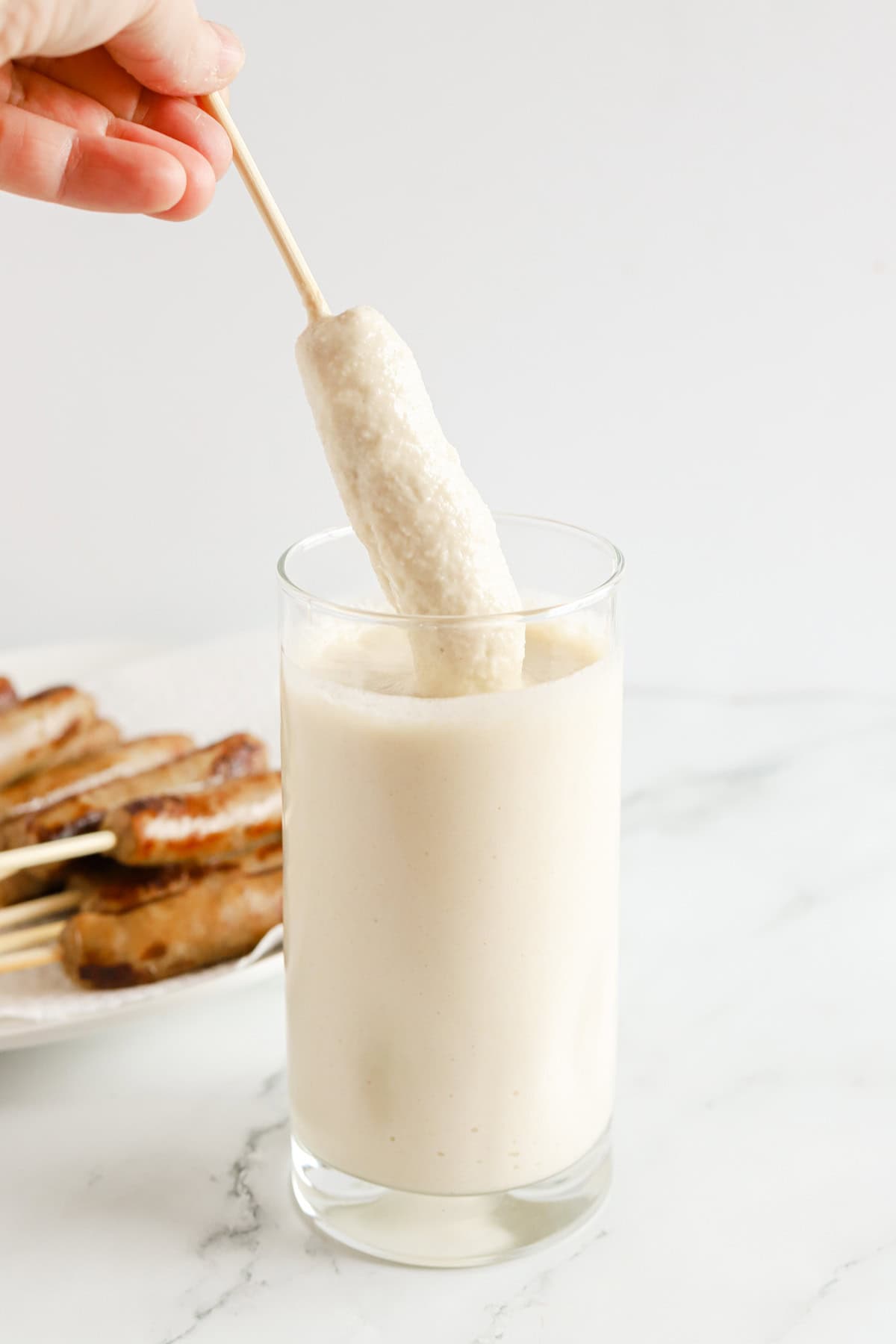 Hand dipping a wooden skewer of sausage into pancake batter in a tall glass.