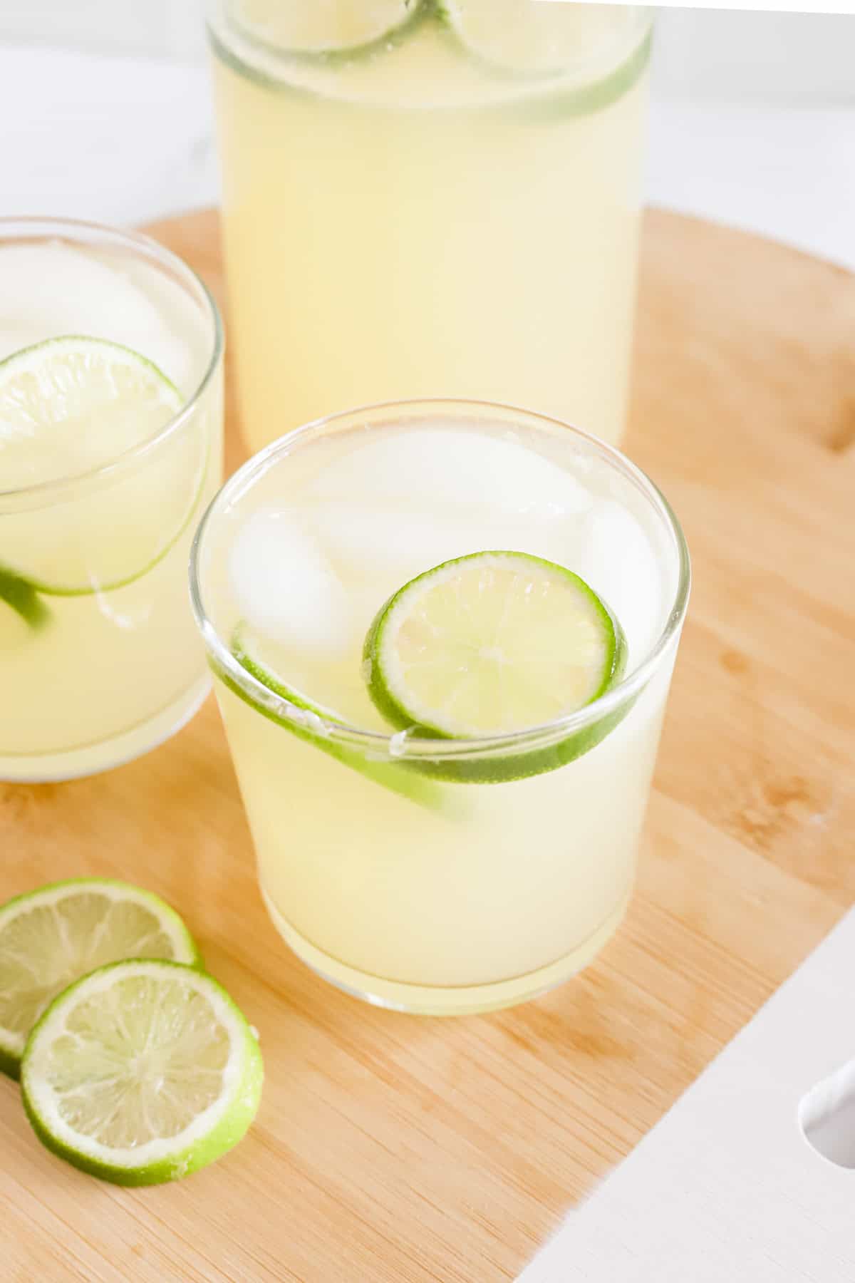 Limeade in two glasses with ice and lime slices on a wooden tray.