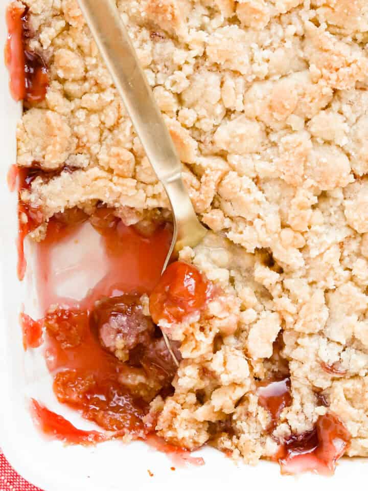 Gold spoon scooping out cherry cobbler that's in a white baking dish with a red napkin underneath the corner.