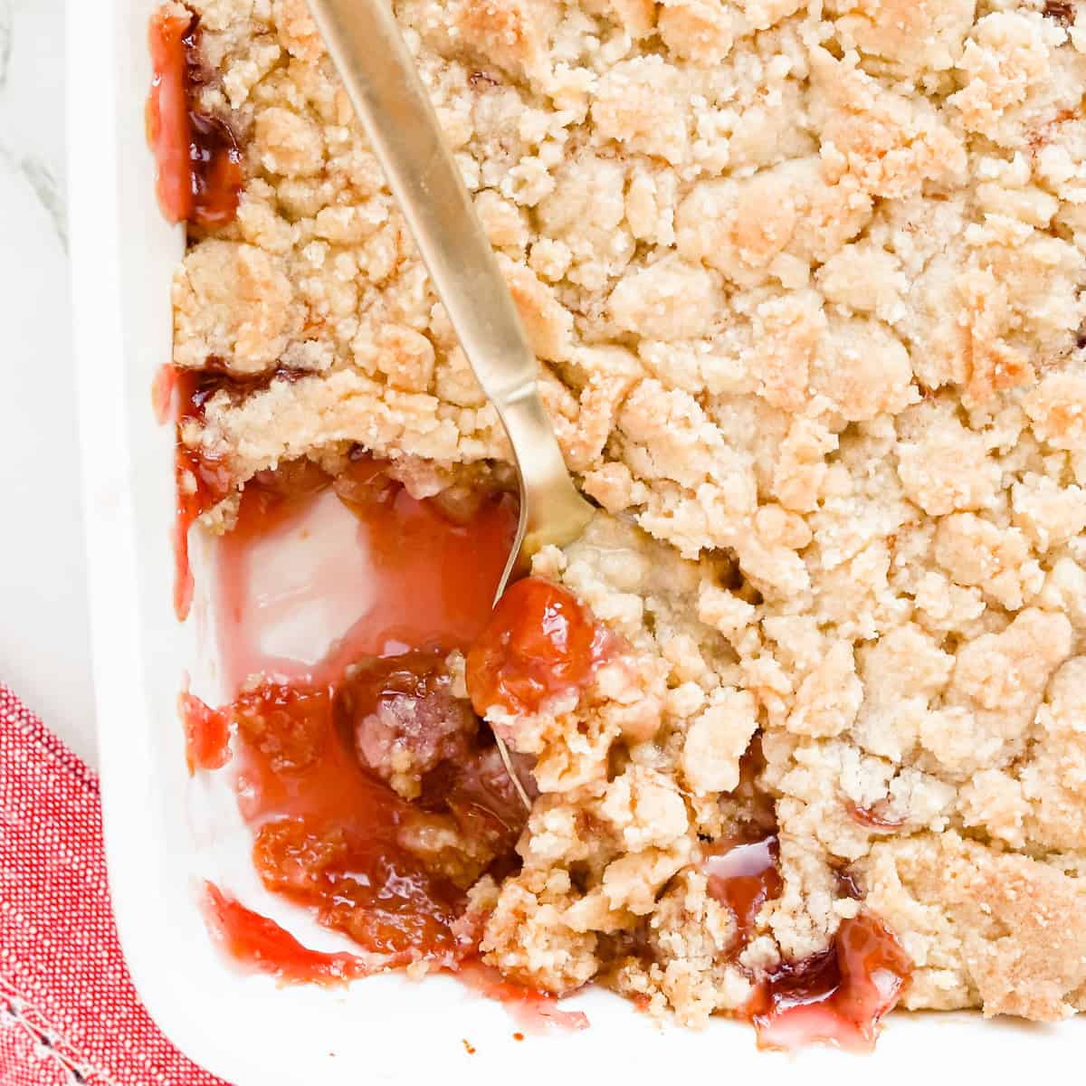 Gold spoon scooping out cherry cobbler that's in a white baking dish with a red napkin underneath the corner.