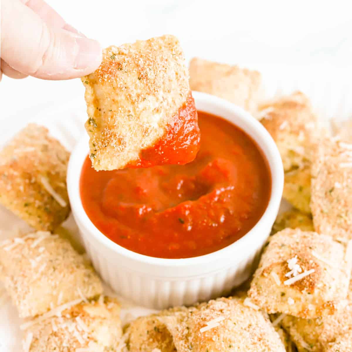 Hand dipping a toasted ravioli in a white bowl of marinara.