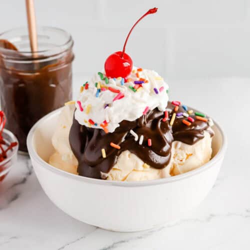 Featured image of a hot fudge sundae in a white bowl with rainbow sprinkles, whipped cream and a cherry on top.