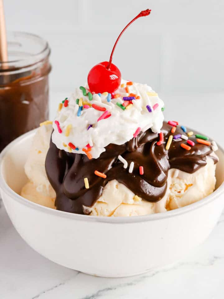 Featured image of a hot fudge sundae in a white bowl with rainbow sprinkles, whipped cream and a cherry on top.