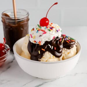 Featured image of a hot fudge sundae in a white bowl with rainbow sprinkles, whipped cream and a cherry on top.