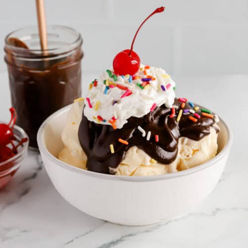 Featured image of a hot fudge sundae in a white bowl with rainbow sprinkles, whipped cream and a cherry on top.