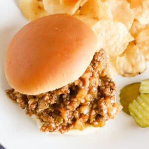 Overhead of a sloppy Joe sandwich with potato chips and pickle slices on a white plate.