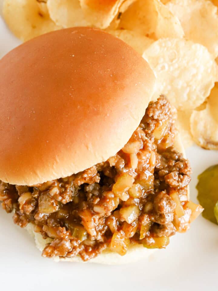 Overhead of a sloppy Joe sandwich with potato chips and pickle slices on a white plate.
