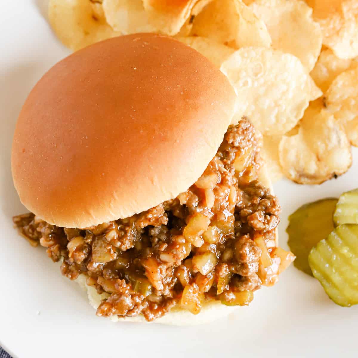 Overhead of a sloppy Joe sandwich with potato chips and pickle slices on a white plate.