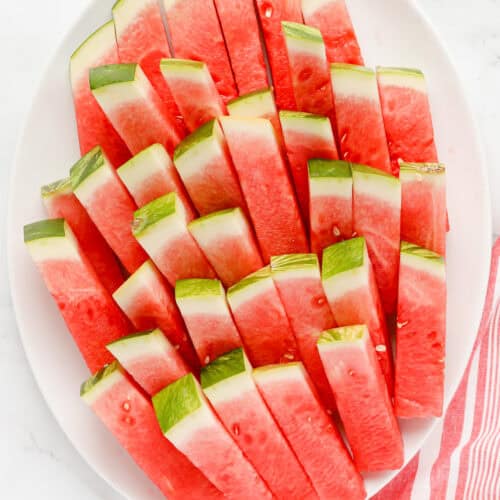 White oval platter of watermelon sticks stacked.