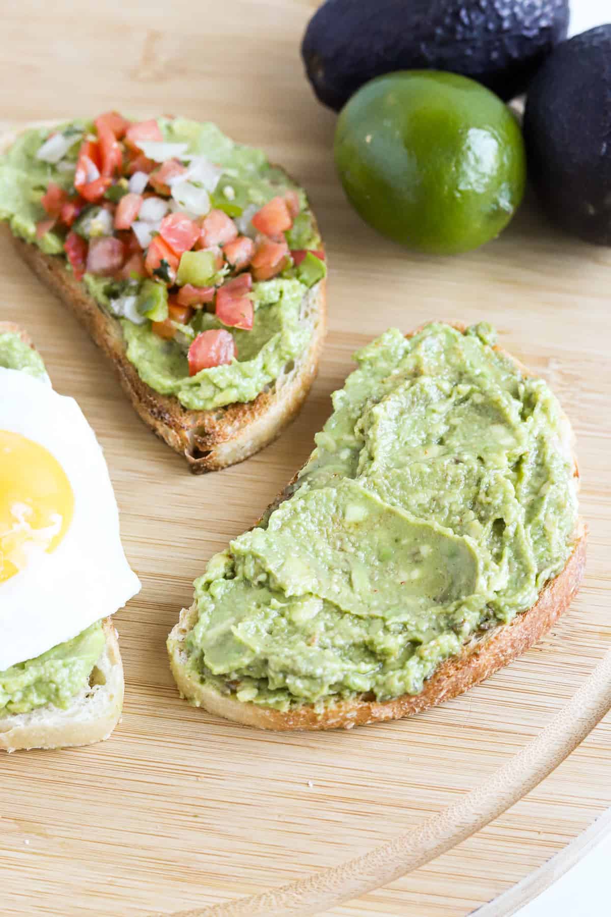 Three slices of guacamole toast on a wooden tray with whole avocados and a lime.