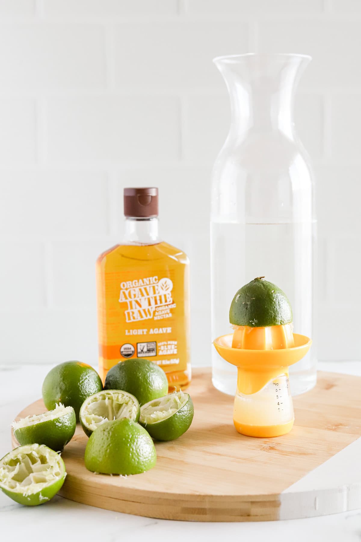 Citrus juicer, carafe of water, juiced limes and bottle of agave nectar on a round wooden tray.