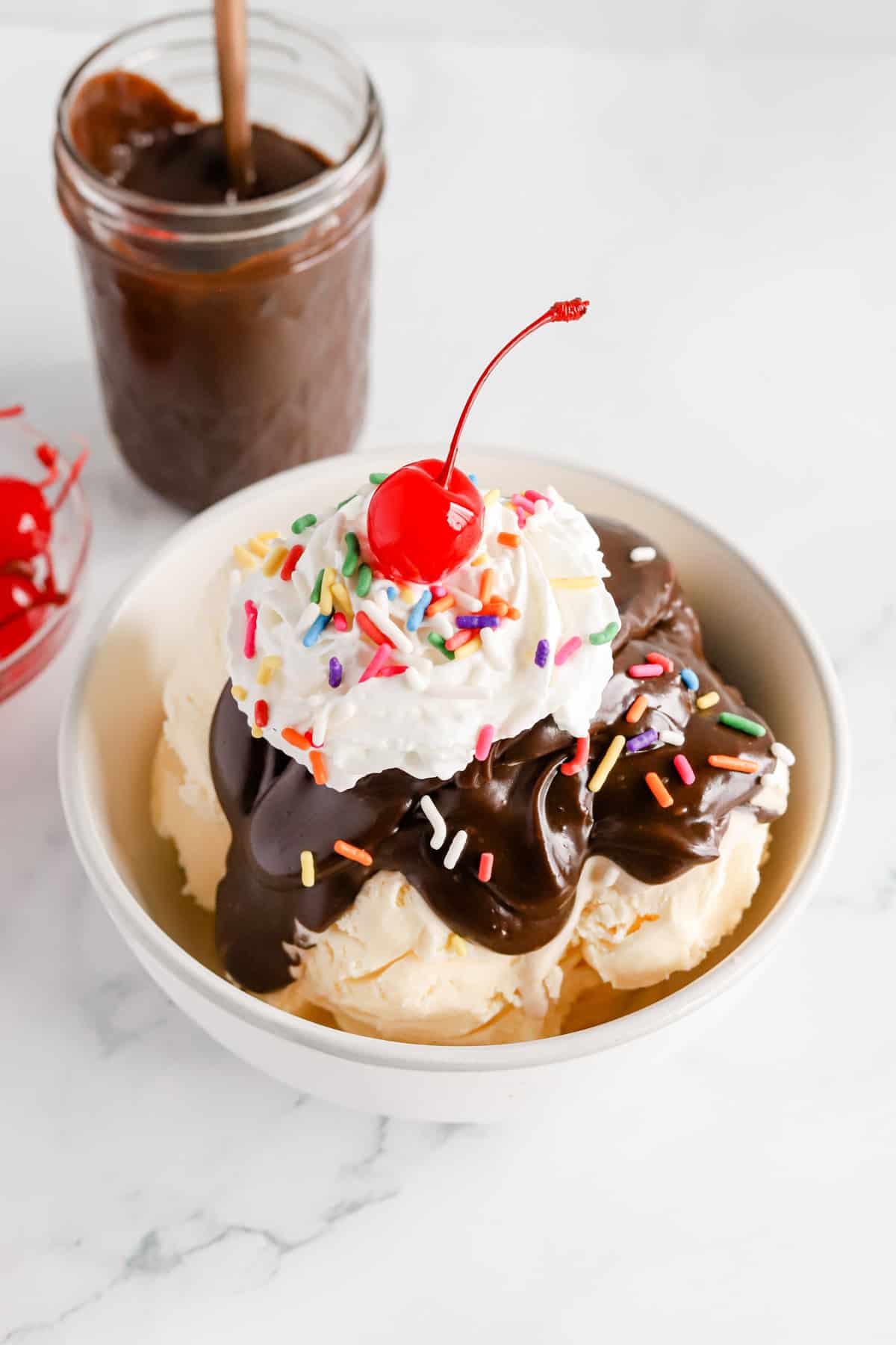 Hot fudge sundae in a white bowl with a jar of hot fudge and cherries in a bowl behind it.