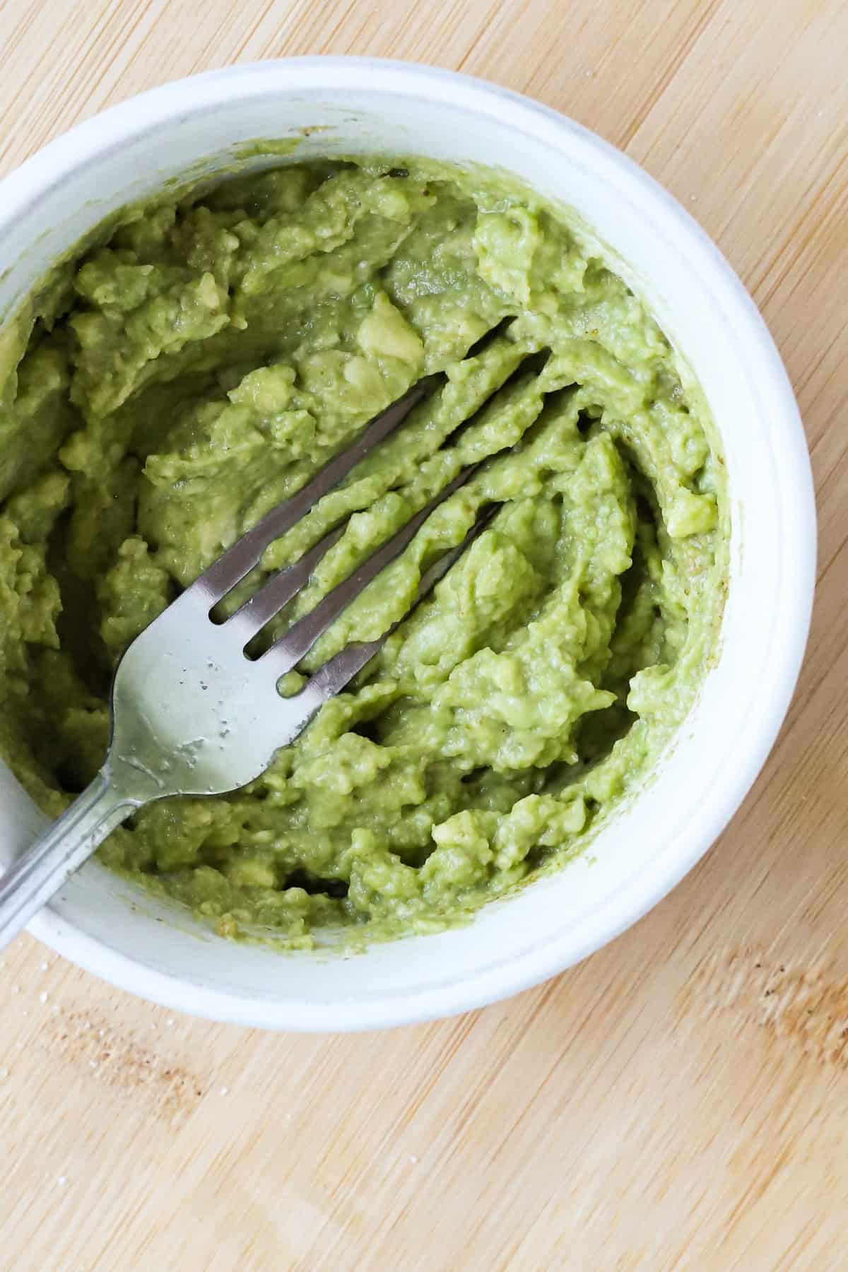 Fork mashing avocado in a white bowl.
