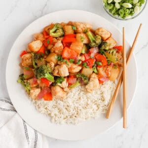 Pineapple chicken stir fry on a white plate over white rice with wooden chopsticks.