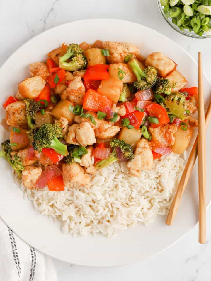Pineapple chicken stir fry on a white plate over white rice with wooden chopsticks.