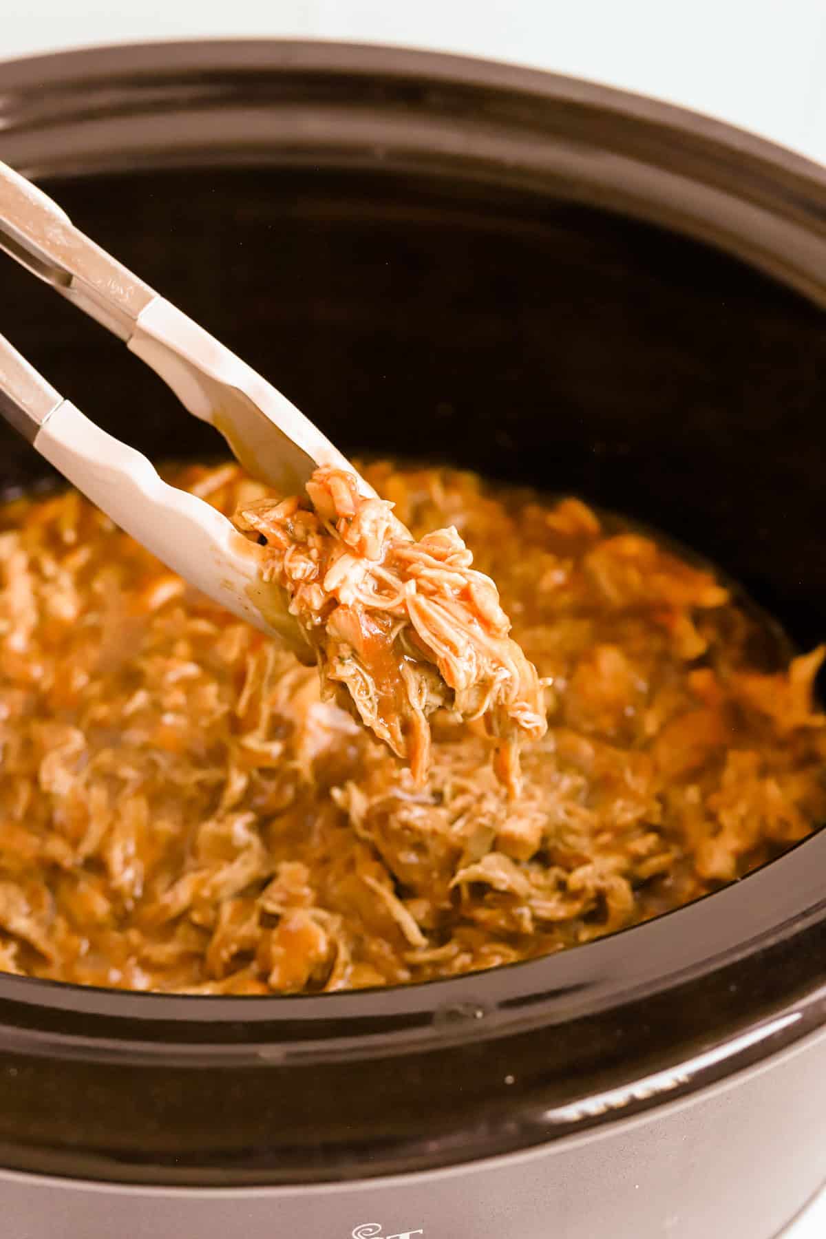 Tongs lifting shredded bbq chicken out of crockpot