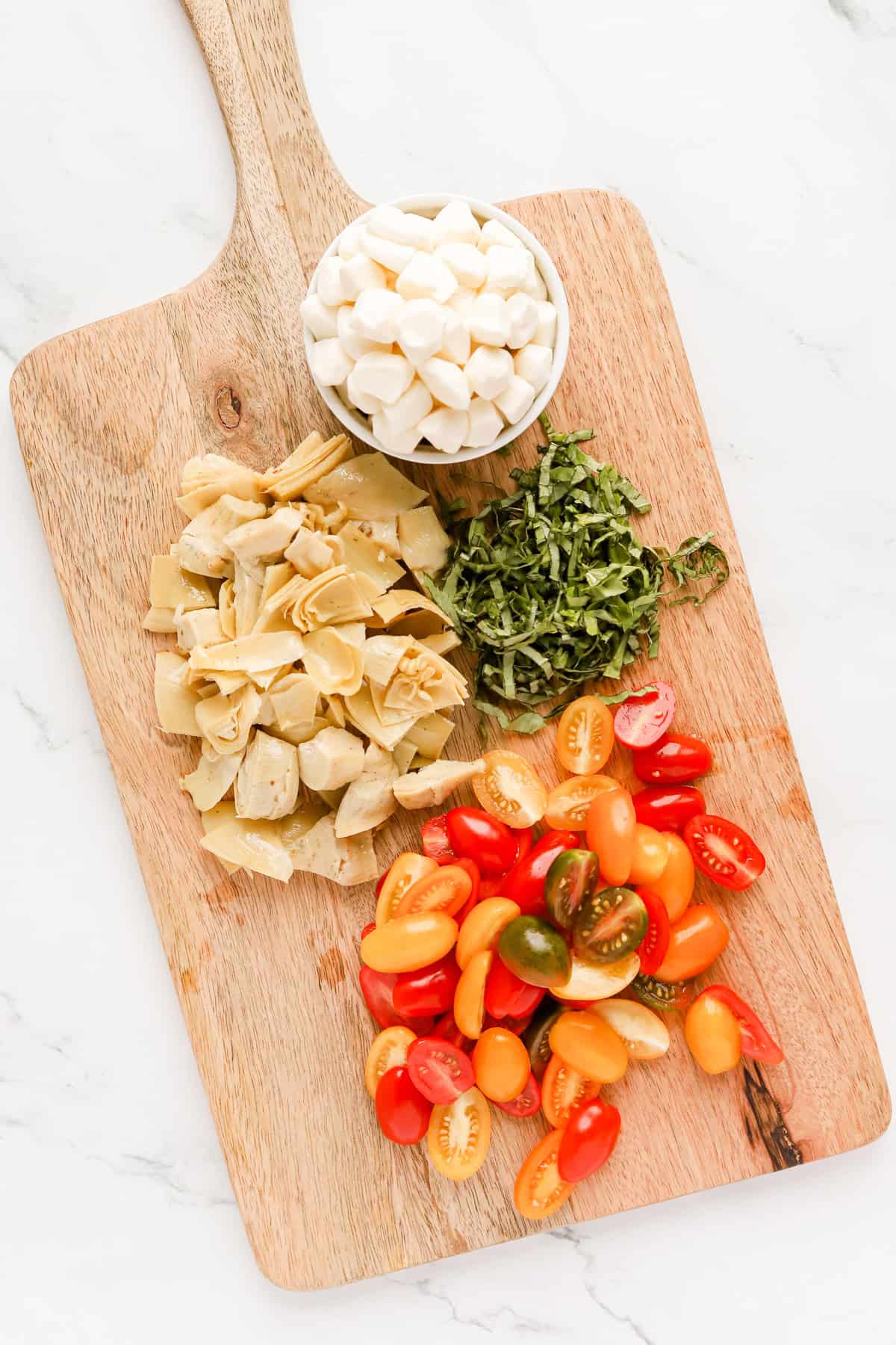 Halved cherry tomatoes, chopped artichokes, chopped basil and mozzarella pearls on a wooden cutting board.