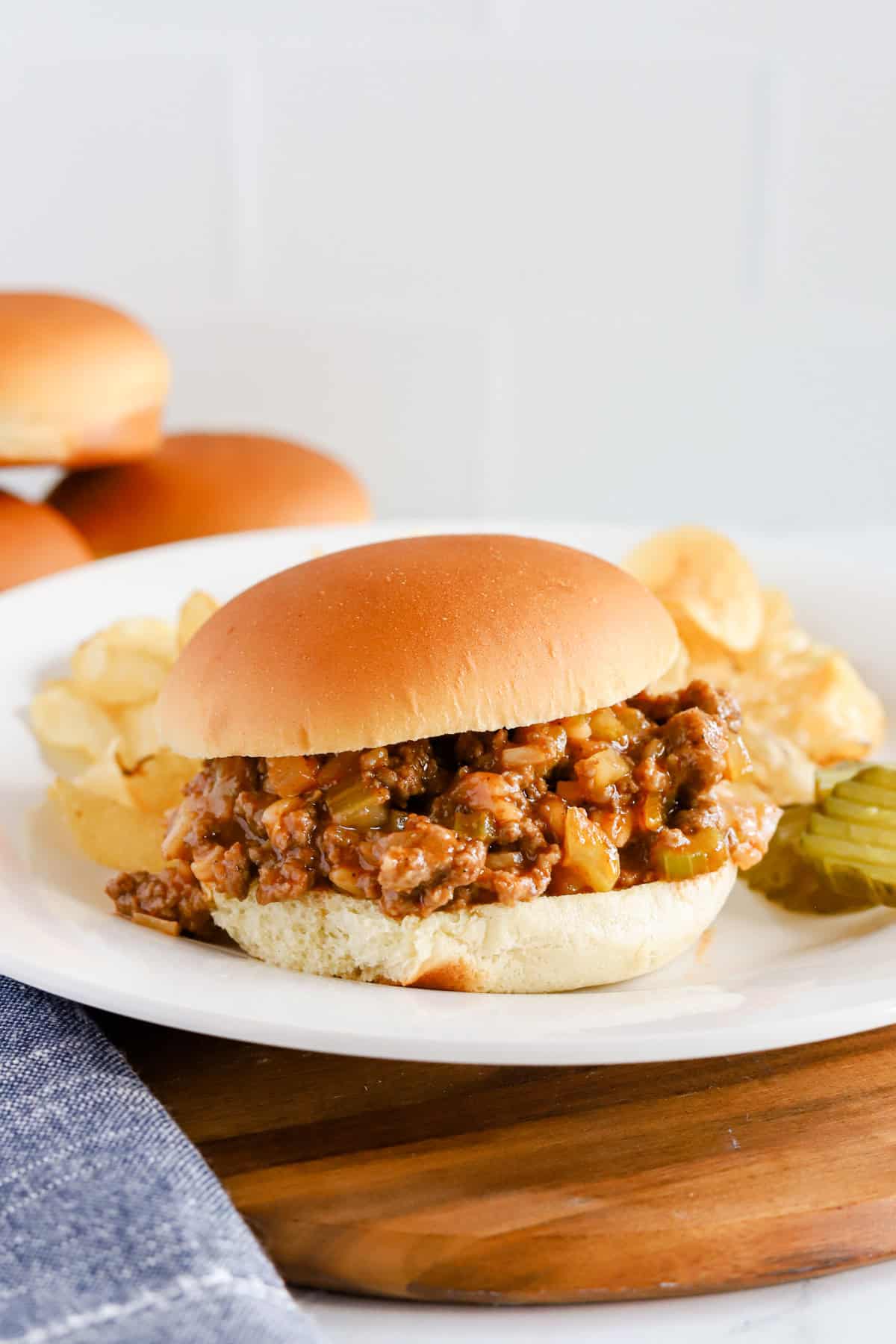 Sloppy Joe on a white plate with potato chips and pickles on a wooden tray.