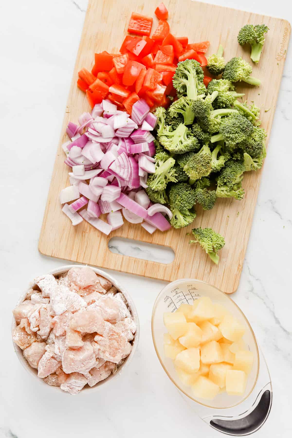 Chopped broccoli, red pepper and red onion a wooden cutting board alongside a bowl of cornstarch coated chicken pieces and clear measuring cup of pineapple chunks.