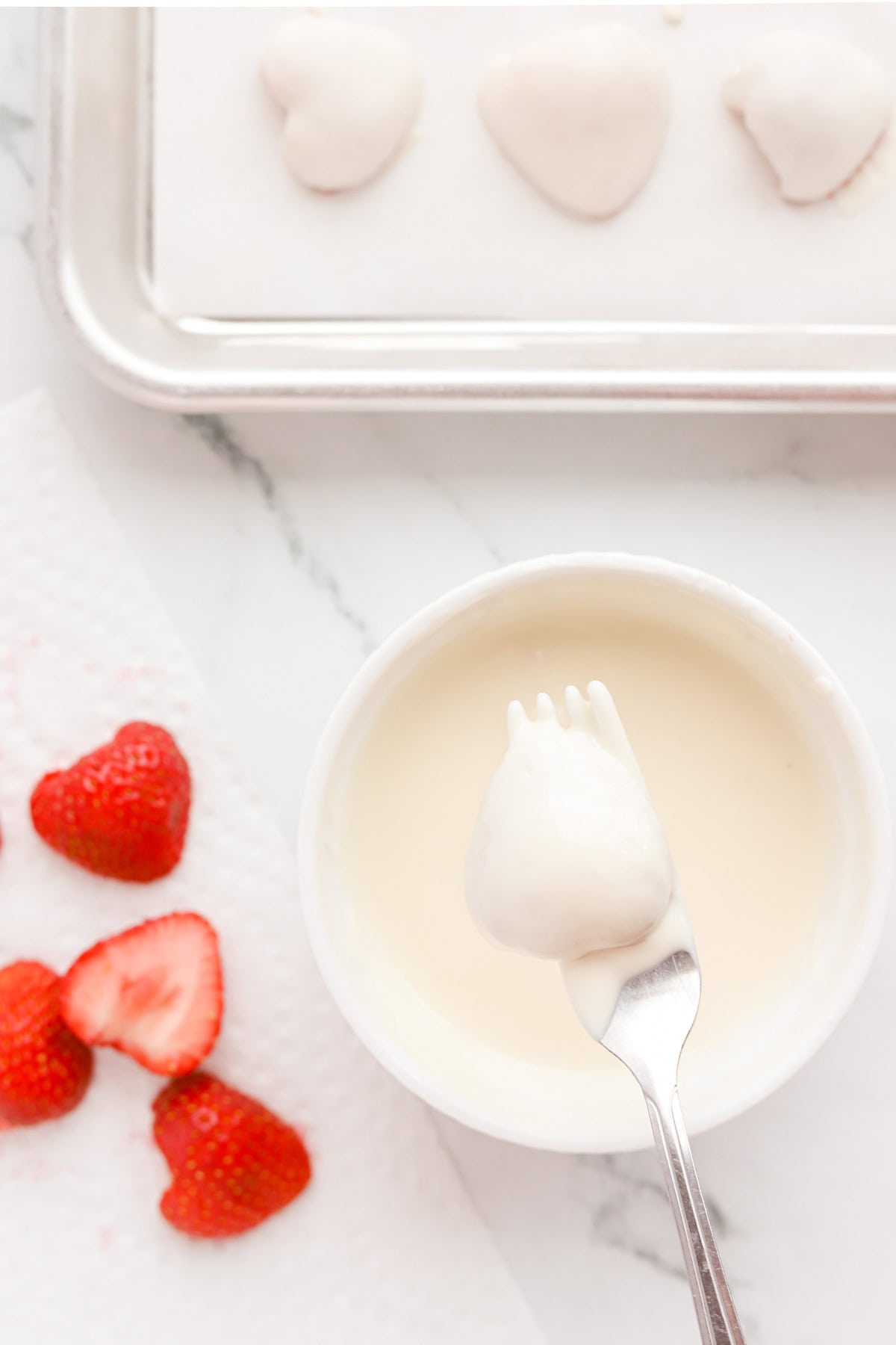 Fork dipping strawberry piece in melted white chocolate. Parchment lined baking sheet with dipped berries above.