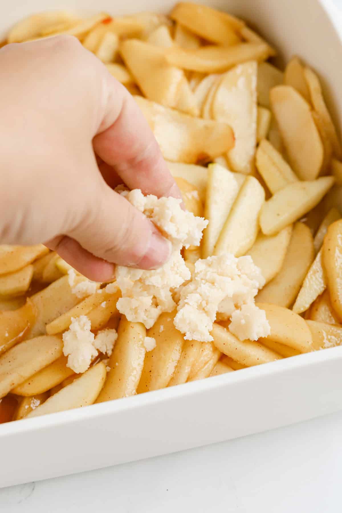 Hand dropping cake mix topping on apple filling in baking dish.
