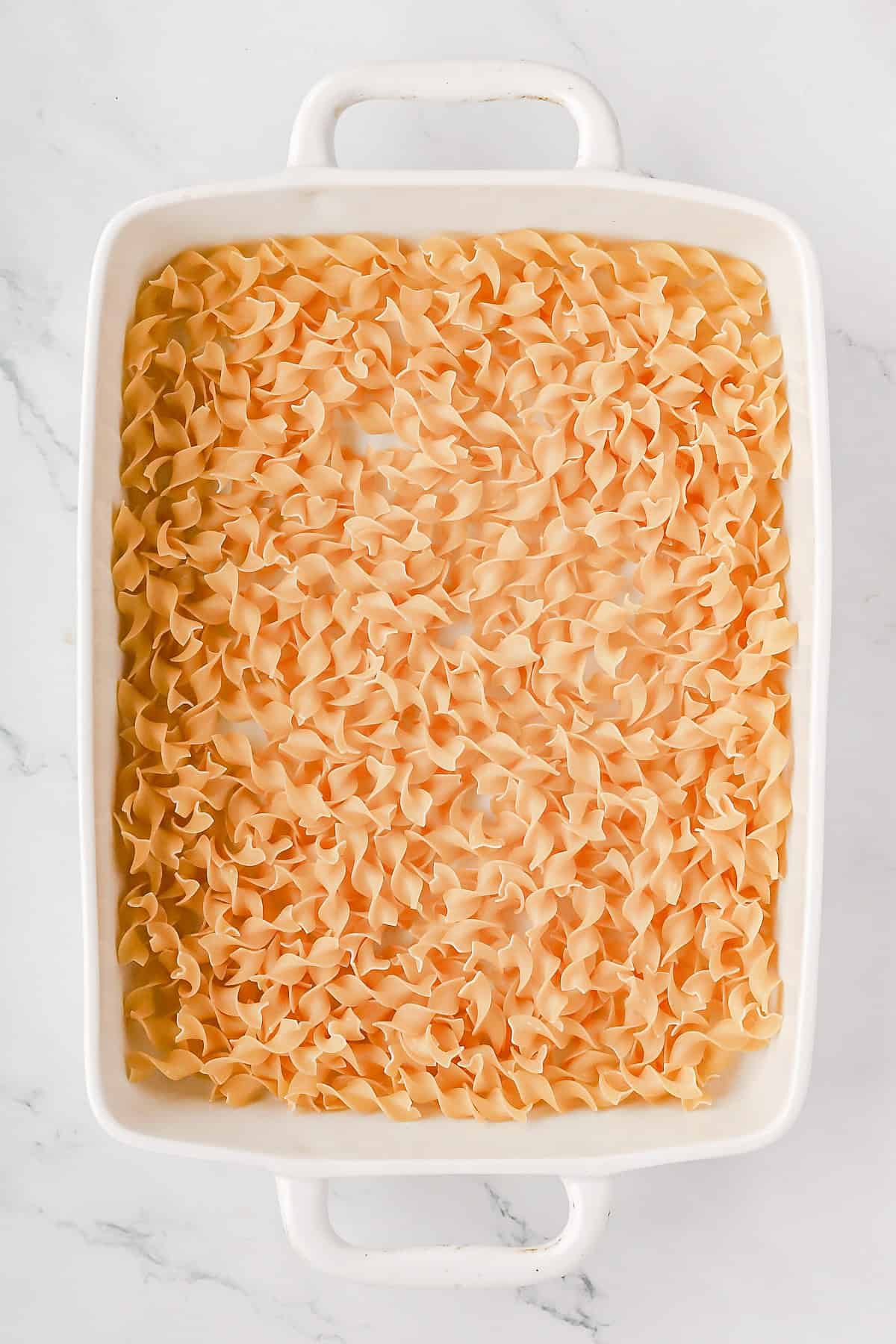 Uncooked wide egg noodles in bottom of white 9x13 baking dish.