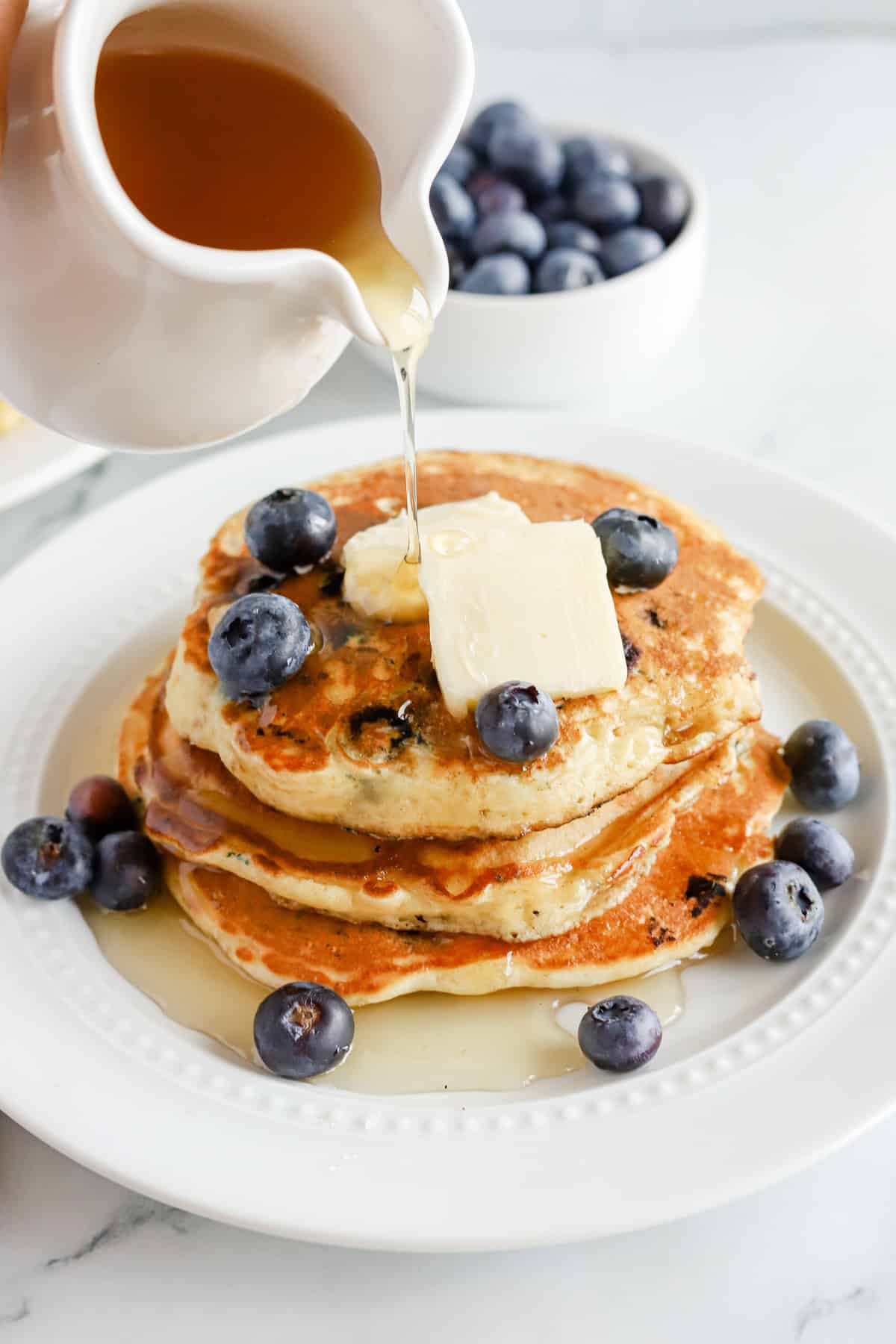 Maple syrup poured over a stack of blueberry pancakes with butter and extra blueberries.