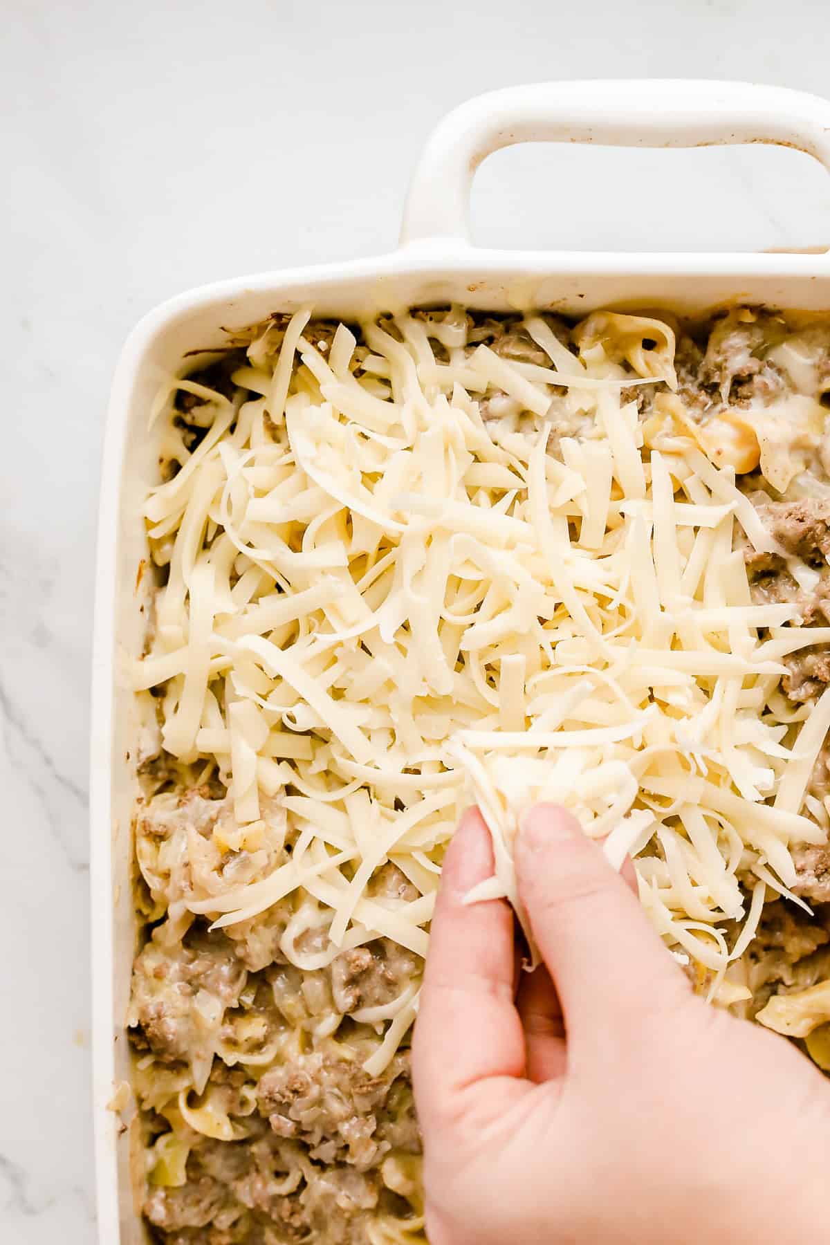 Hand sprinkling shredded Swiss cheese on top of casserole in white baking dish.